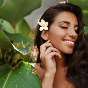 charming woman with white flower dark hair smiles sweetly with closed eyes among tropical tree with large leaves